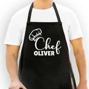 以下が含まれることがあります： 白い文字で「Chef OLIVER」とシェフの帽子グラフィックがプリントされた黒いエプロン。エプロンにはビブとネックストラップが付いています。エプロンは白いTシャツの上に着用されています。