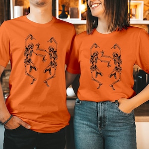 May include: A bright orange short-sleeved t-shirt featuring a black graphic print of two skeletons in a dancing pose. The skeletons are positioned side-by-side, with arms and legs extended in dynamic motion, centred on the front of the shirt.