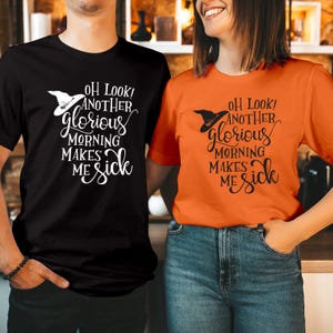 Op de afbeelding: Een zwart t-shirt en een oranje t-shirt, beide voorzien van een afbeelding van een heksenhoed en de tekst "OH LOOK! ANOTHER glorious MORNING MAKES ME sick" in een contrasterende kleur. Het zwarte shirt heeft witte tekst, het oranje shirt zwarte.