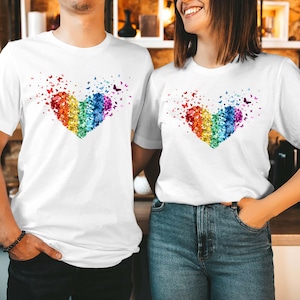 May include: Two white t-shirts featuring a heart design composed of colourful butterflies. The heart is a rainbow of colours, including red, orange, yellow, green, blue, and purple. The shirts are worn by two people.