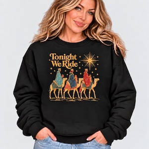 May include: A black crewneck sweatshirt featuring a graphic print of three figures wearing crowns and robes riding camels. Above the figures, the text "Tonight We Ride" is printed in gold, accompanied by a large star and smaller star accents.