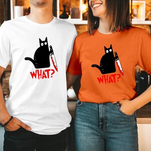 Może przedstawiać: Biały t-shirt i pomarańczowy t-shirt, każdy z grafiką czarnego kota trzymającego nóż z czerwonymi plamami krwi na ostrzu. Pod kotem na obu ubraniach nadrukowano słowo WHAT? pogrubionymi czerwonymi wielkimi literami.