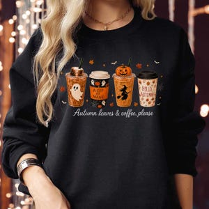 May include: A black long-sleeve sweatshirt featuring a graphic of four coffee cups with autumn and Halloween themes. The cups display a ghost, the text A CUP OF MAGIC, a witch silhouette with a pumpkin, and the text HELLO, AUTUMN! with leaves.