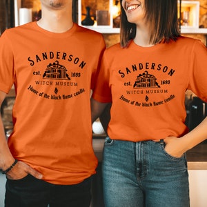May include: An orange short-sleeved t-shirt featuring a black graphic print of a house with the text Sanderson est. 1693 Witch Museum Home of the black flame candle. The design includes a small flame icon centered below the museum name.