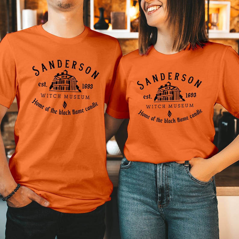 May include: An orange short-sleeved t-shirt featuring a black graphic print of a house with the text Sanderson est. 1693 Witch Museum Home of the black flame candle. The design includes a small flame icon centered below the museum name.