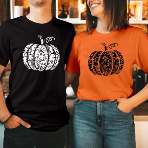 May include: A black t-shirt with a white pumpkin graphic featuring an intricate floral scrollwork pattern and an orange t-shirt with a black pumpkin graphic featuring the same intricate floral scrollwork pattern, both with short sleeves.
