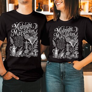 May include: A black t-shirt featuring a white graphic print with the text Midnight Margaritas. The design includes a margarita glass, a teapot, a toad wearing a witch hat, a mushroom, and a recipe for a cocktail, all surrounded by stars and moon.