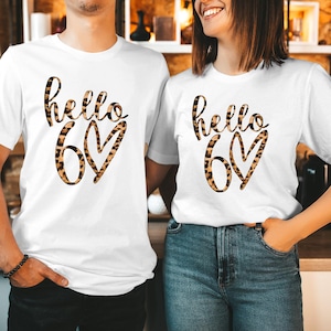 May include: White t-shirts with the text "hello 60" in a leopard print design. The number 60 is inside a heart shape. The shirts are worn by two people, one with a bracelet and the other with their hand in their pocket.