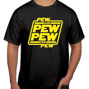 May include: Black t-shirt featuring a yellow graphic with the text "PEW PEW PEW" in a bold, stylized font. The design is enclosed within a yellow rectangular border. The shirt is short-sleeved and likely made of cotton.