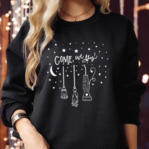 May include: A black long-sleeve sweatshirt featuring a white graphic print of stars, a crescent moon, a broom, a mop, and a vacuum cleaner. The text "COME, we fly!" is printed in white script above the illustrations of the cleaning tools.