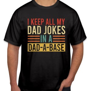 May include: Black crew neck t-shirt featuring the text "I KEEP ALL MY DAD JOKES IN A DAD-A-BASE" in a retro style. The text is in red, beige, teal, and orange. The design has a distressed look.