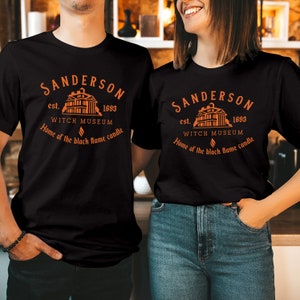 May include: A black t-shirt featuring an orange graphic print of a building with the text Sanderson est. 1693 Witch Museum Home of the black flame candle. The design includes a small flame icon centered below the museum name and above the text.