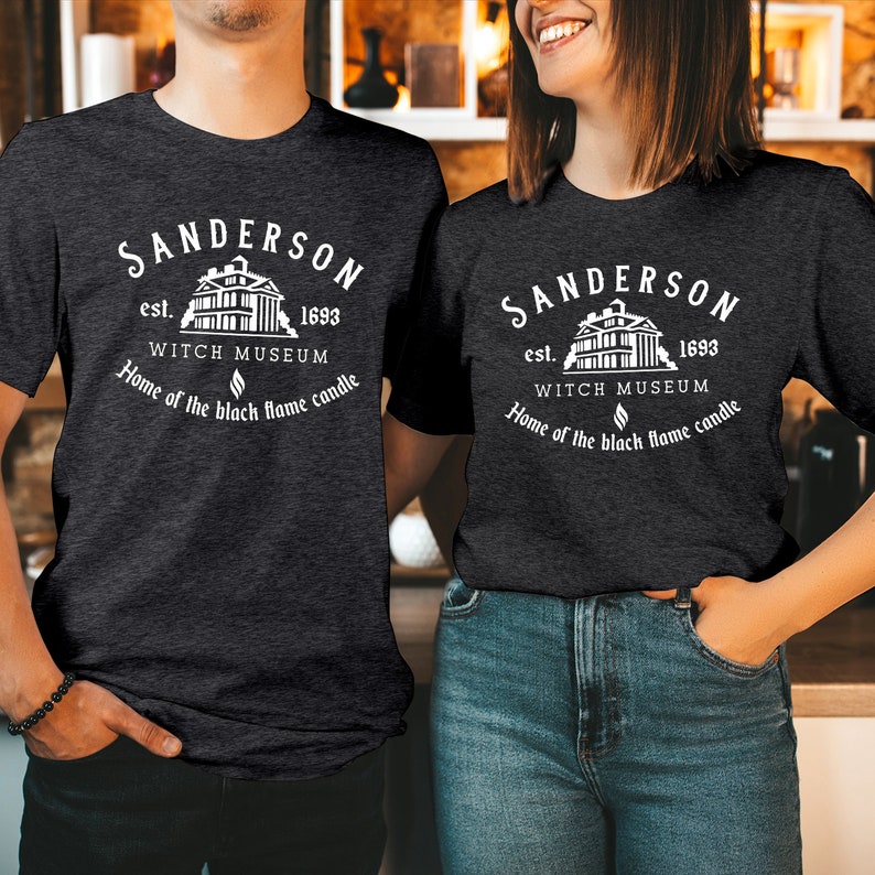 May include: A dark charcoal gray short-sleeve t-shirt featuring a white graphic print that reads Sanderson Witch Museum est. 1693, Home of the black flame candle, accompanied by an illustration of a house with columns and a small flame icon.