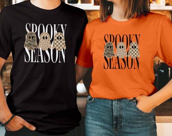 Op de afbeelding: Een zwart t-shirt en een oranje t-shirt, beide voorzien van de tekst "SPOOKY SEASON" in zwarte of witte letters. Elk shirt toont op de borst een afbeelding van drie geesten met camouflage-, luipaard- en ruitpatronen.