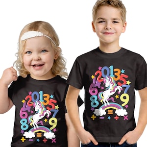 May include: Two children wearing black t-shirts with a colourful unicorn design. The shirts feature a unicorn standing on a rainbow, surrounded by numbers and stars. The design includes the numbers 2, 0, 1, 3, 5, 6, 7, 8, and 9.