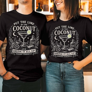 May include: A black t-shirt featuring a white graphic illustration of a margarita glass, a bottle labeled Witches Spirits, and citrus slices. The text reads: PUT THE LIME IN THE COCONUT AND DRINK IT ALL UP MIDNIGHT MARGARITAS in a decorative font.