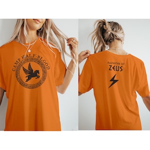 May include: Orange t-shirt with black graphic text and design. The front of the shirt features the words "CAMP HALF-BLOOD" around a circular design with a winged horse. The back of the shirt says "DAUGHTER OF ZEUS" with a lightning bolt.