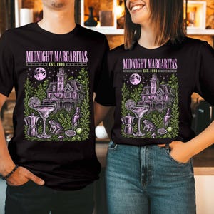 May include: A black t-shirt featuring a graphic print with the text "MIDNIGHT MARGARITAS EST. 1998". The design includes a large house, a full moon, a margarita glass, a frog, a coffee pot, a bottle, and lime slices in shades of purple and green.