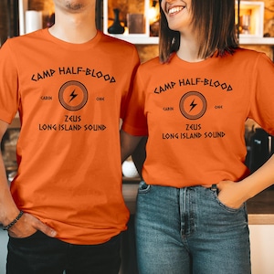Puede incluir: Dos camisetas naranjas con el texto "CAMP HALF-BLOOD" en negro. Debajo hay un círculo con un rayo y las palabras "CABIN" y "ONE". Debajo está "ZEUS LONG ISLAND SOUND".