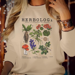 May include: Cream-colored sweatshirt featuring a botanical illustration with the text "HERBOLOGY SCHOOL OF WITCHCRAFT AND WIZARDRY." The graphic includes drawings of various plants, including a mandrake, toadstools, and gillyweed.