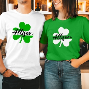 May include: Two short-sleeved t-shirts, one white and one green, each displaying a shamrock design with the name "Allison" in black script. The shamrock is a symbol of St. Patrick's Day. The shirts are likely made of cotton.