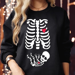 May include: A black long-sleeve sweatshirt featuring a white graphic of a human rib cage with a small red heart on the left side. Below the rib cage is a white graphic of a small skeleton with heart-shaped eye sockets and a tiny red heart.