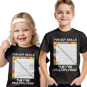 以下が含まれることがあります： 掛け算表のデザインが施された黒いTシャツを着た2人の子供たち。シャツには白い文字で「I'VE GOT SKILLS」と「THEY'RE MULTIPLYING！」と書かれています。一人の子供は白いヘアバンドをしています。