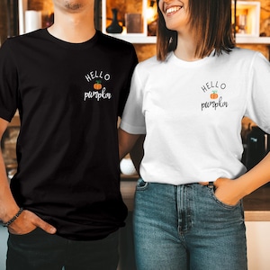 May include: A black short-sleeved t-shirt and a white short-sleeved t-shirt, each featuring a small graphic on the left chest area. The graphic consists of the text "HELLO pumpkin" in black lettering with a small orange pumpkin illustration.