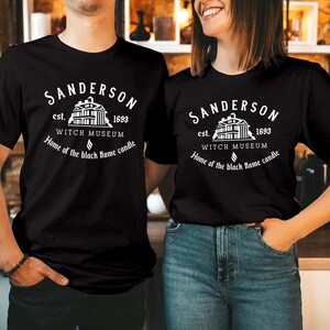 May include: A black t-shirt featuring a white graphic print that reads Sanderson Witch Museum, est. 1693, Home of the black flame candle, accompanied by an illustration of a building and a small flame icon centered on the chest area.