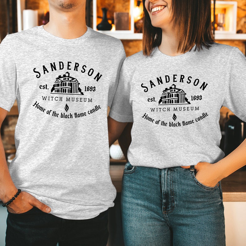 May include: A light gray t-shirt featuring a black graphic of a building with the text: SANDERSON est. 1693 WITCH MUSEUM Home of the black flame candle. The design includes a small flame icon centered below the museum name and above the final line.