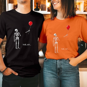 May include: A black t-shirt and an orange t-shirt, each featuring a graphic of a skeleton looking up at a red balloon held by a detached skeletal hand, with the text "Well, shit..." printed in white below the balloon string.