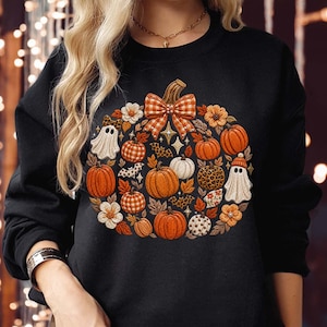 May include: A black long-sleeve sweatshirt featuring a large central graphic of a pumpkin shape composed of smaller pumpkins, white ghost figures, orange and white flowers, autumn leaves, and a plaid bow, all arranged in a circular pattern.