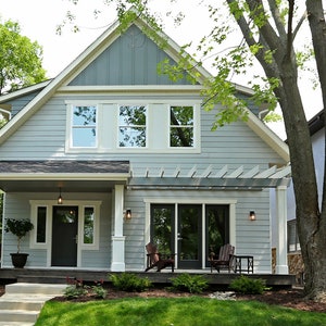 May include: A two-storey grey house with white trim and a white pergola over the front porch. The house has a front porch with a black door and two white columns. There are two chairs on the porch and a small tree in a pot.