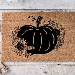 May include: A brown coir doormat with a black silhouette of a pumpkin with a stem and leaves. The pumpkin is surrounded by black silhouettes of sunflowers and other flowers.