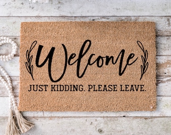 May include: A brown coir doormat with the text "Welcome" in a black cursive font with leafy accents on either side. Below the text is the phrase "Just kidding. Please leave."