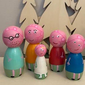 May include: Six wooden peg dolls, each with a pink head and a body painted in different colours. The dolls are designed to resemble a cartoon family, with painted facial features and clothing.