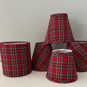 May include: A collection of five fabric lampshades featuring a red, green, yellow, and blue plaid pattern. The set includes one cylindrical shade and four tapered shades of varying sizes, all with finished edges and a white interior lining.