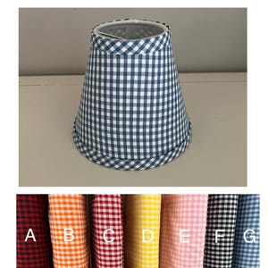 May include: A blue and white gingham lampshade with a flared shape. The lampshade has a white interior and a matching gingham trim at the top and bottom. Below the lampshade are fabric swatches in various colors, including red, orange, and pink.