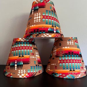 May include: Three conical lamp shades featuring a tan background with a repeating geometric pattern in vibrant shades of turquoise, red, orange, black, and white. The shades are arranged in a pyramid formation with one on top and two below.