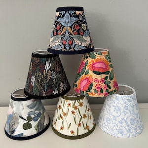 May include: A collection of six conical lamp shades featuring diverse patterns. The top shade has a dark blue background with birds and strawberries. Below are shades with botanical prints, abstract brushstrokes, yellow daffodils, and blue floral motifs.