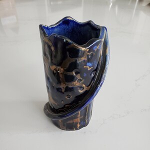 May include: A handcrafted ceramic vase with a dark blue glaze and gold accents. The vase has a unique, wavy rim and a decorative, curved handle. The exterior features a textured surface with small, circular indentations. The vase is approximately 7 inches tall.