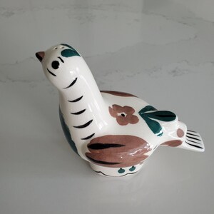 May include: A ceramic bird-shaped salt shaker. The bird is primarily white with brown, teal, and black painted details. The design includes floral accents and a black beak. The salt shaker has a small opening on top for dispensing salt.