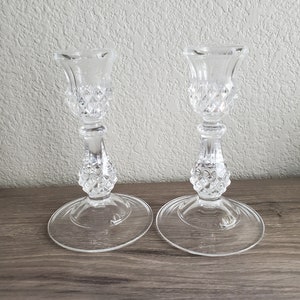 May include: Two clear glass candle holders with diamond-cut detailing. Each holder has a round base, a slender stem, and a flared top. The candle holders are displayed on a wooden surface against a textured wall.