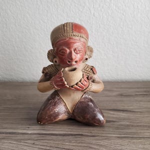 May include: A terracotta figurine of a kneeling figure holding a small vessel. The figure has a red face and head with detailed features, wearing a headdress. The body is tan and brown, with the figure's hands and knees visible.