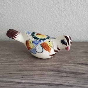 May include: A ceramic bird figurine with a cream-colored body and colorful floral designs. The bird has brown and white striped details on its head and tail, with a pink beak and black eyes. The figurine is placed on a wooden surface.