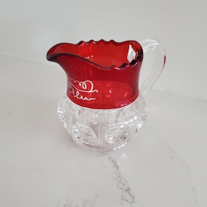 May include: A small, clear glass pitcher with a ruby red rim and handle. The pitcher has a decorative, cut-glass design on the lower portion. White script is visible on the red rim. The pitcher is likely for serving liquids.