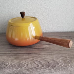 May include: A yellow and orange enameled saucepan with a wooden handle and lid. The pot has a rounded shape and a dark brown wooden knob on the lid. The handle is also made of wood. The pot is sitting on a wooden surface.