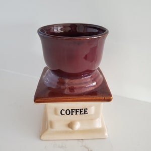 May include: A ceramic coffee grinder with a dark maroon cup-shaped top and a cream-colored base. The base has the word "COFFEE" printed in black. The grinder is a decorative kitchen item.