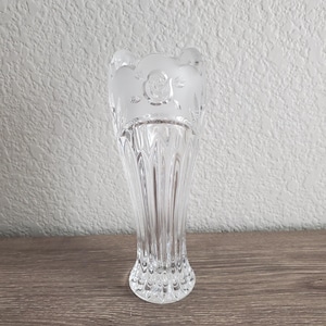 May include: Clear glass vase with a fluted body and a frosted floral design at the top. The vase has a flared opening and a detailed base. The background is a textured white wall and a wooden surface.