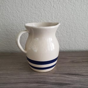 May include: Cream-colored ceramic pitcher with a rounded body and a handle. Two navy blue stripes encircle the base. The pitcher has a small spout for pouring. The background is a textured white wall and a wooden surface.